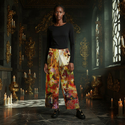 A model stands in a dark, ornate hall with candles and gold detailing, wearing a black long-sleeve top and wide-leg pants with a colorful abstract print in red, yellow, orange, and cream.