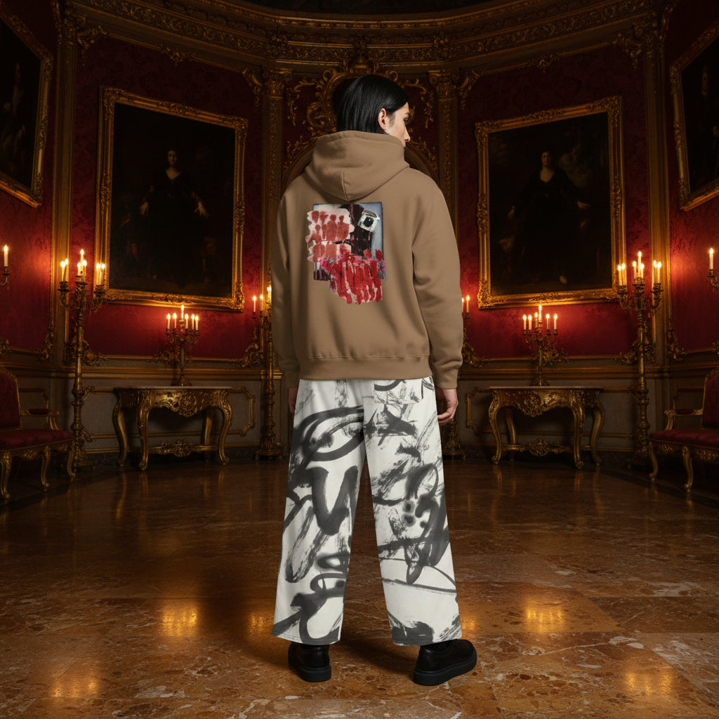 A person stands in a grand, ornate room with red walls, gold-framed portraits, and candelabras, wearing a tan hoodie with a large red and black graphic on the back and white wide-leg pants with black abstract brushstroke patterns.
