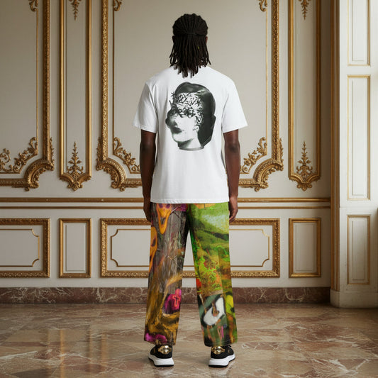 A man stands with his back to the camera in an ornate room with gold-trimmed cream walls and marble floors, wearing a white T-shirt with a large black-and-white portrait graphic on the back and colorful painterly pants.