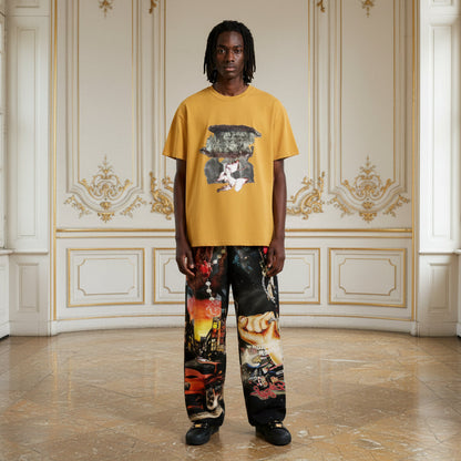 A model stands in a grand, ornate room wearing a mustard yellow graphic t-shirt and wide-leg pants featuring a complex collage print of cars, hands, and abstract art.
