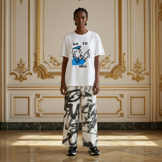 A model stands in a grand, ornate room with gold-trimmed cream walls, wearing a white graphic t-shirt with a blue and black cartoon sailor design and the text 'GO TO HELL', paired with wide-leg white pants featuring a black and gray abstract brushstroke pattern.